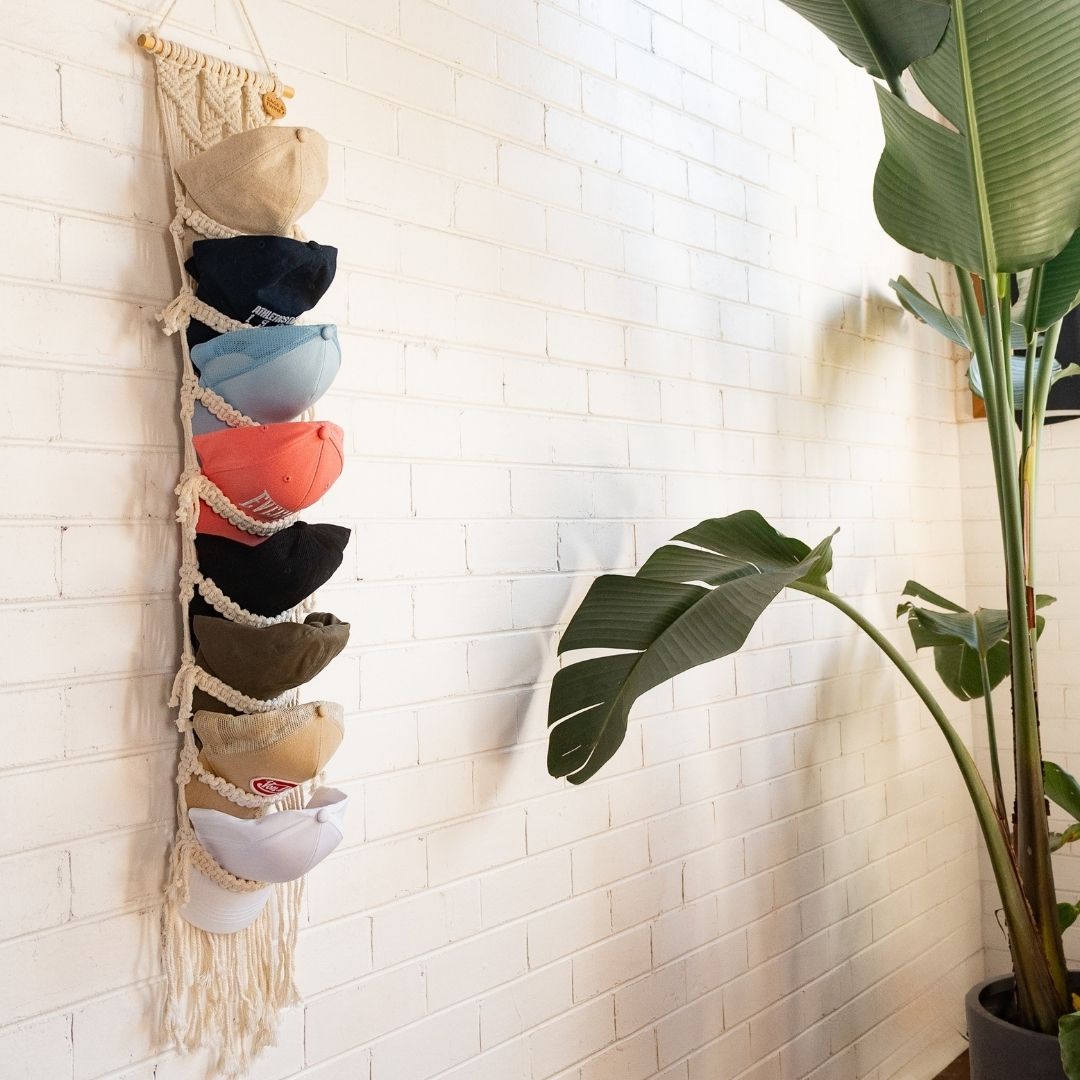 Macrame hat rack with colorful hats against a white brick wall with a plant in the corner by Sage and Twine.