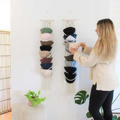 macrame cap hangers hang on a white wall in a home, with a woman placing a baseball cap into the macrame hanging. The macrame pieces hold a dozen baseball caps of many colours and the home features green leafy plants