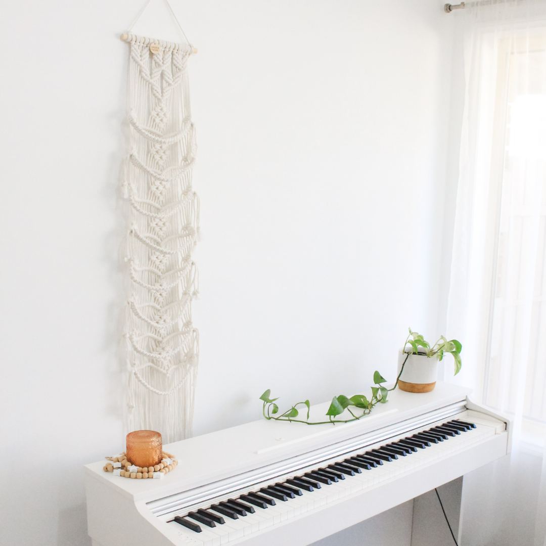 Macrame Cap Hanger made from natural colour cotton hangs above a piano. The room is bright and white and the macrame cap hanger is empty, showing off the macrame details of the hanging