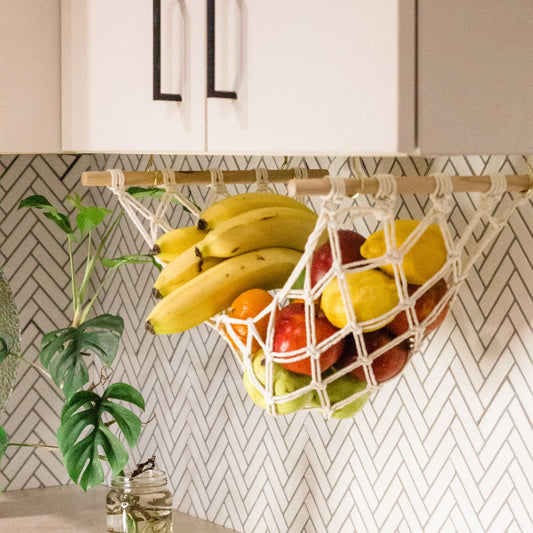 A macrame fruit hammock holding bananas apples lemons oranges and pears hanging from under a kitchen cabinet. handmade by Sage and twine . Australian Owned Business. Mini monstera plant in background