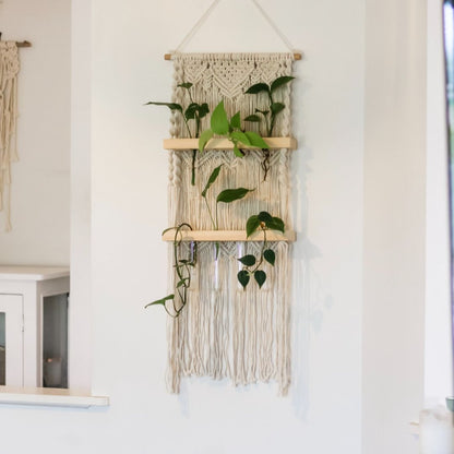 A macrame plant propagation station. The Macrame wall hanging has a wooden dowel at the top and two wooden shelves. Test tubes hang in holes in the shelves and are being used as a propagation stations for greenery cuttings such as pothos, philodendron, and more plants. There is a beautiful pattern knotted into the cotton cord at the top, with a downwards arrow design. this repeats twice behind the shelves. At the bottom of the last shelf there are long tassels hanging from the wood.