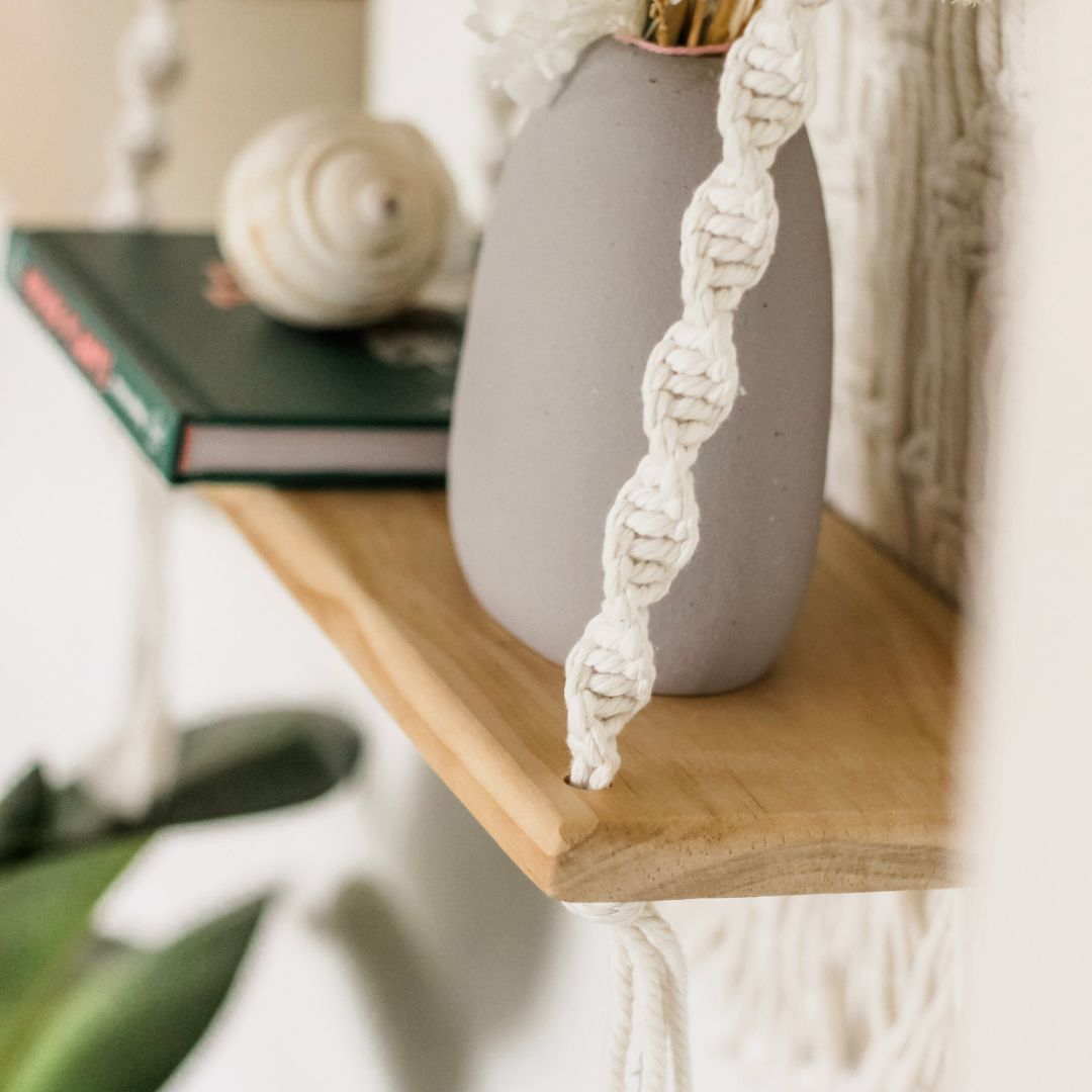 close up of the high quality wooden platform used in the Macrame shelf made in Australia by the brand sage and twine. this image shows the pot plant and "house plants" book and a boho shell.