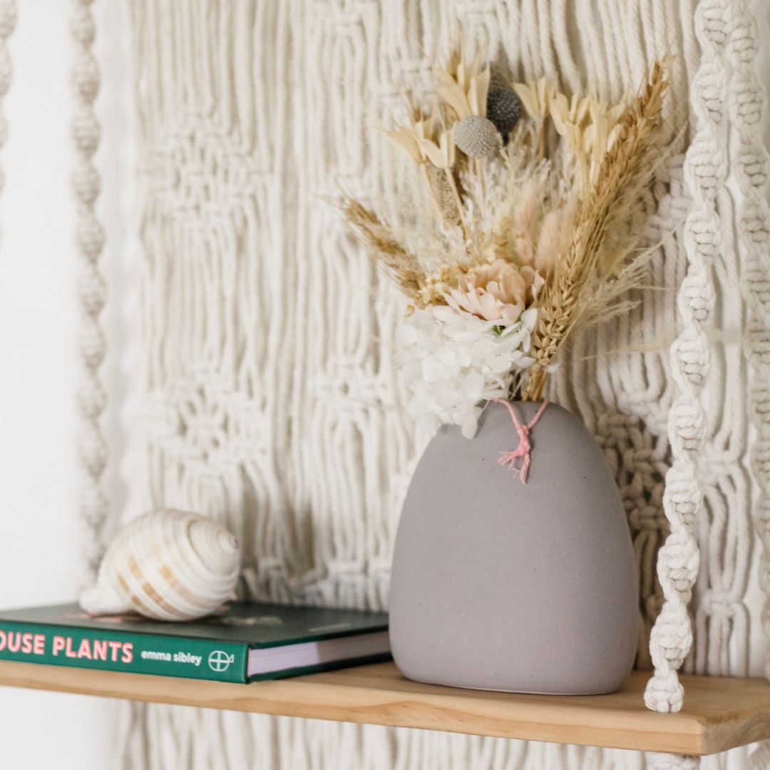 a close up of a macrame shelf and boho decor items on the top of the shelf. there is a shell, book and some boho grass.