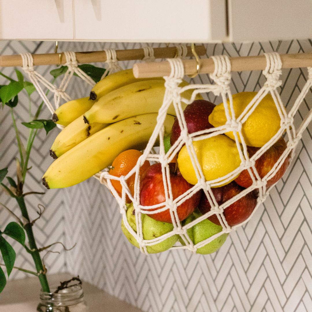 close up of a macrame hammock holding bananas apples lemons oranges and pears hanging from a kitchen cabinet. handmade by Sage and twine . Australian Owned Made with cotton cord and a wooden dowel.