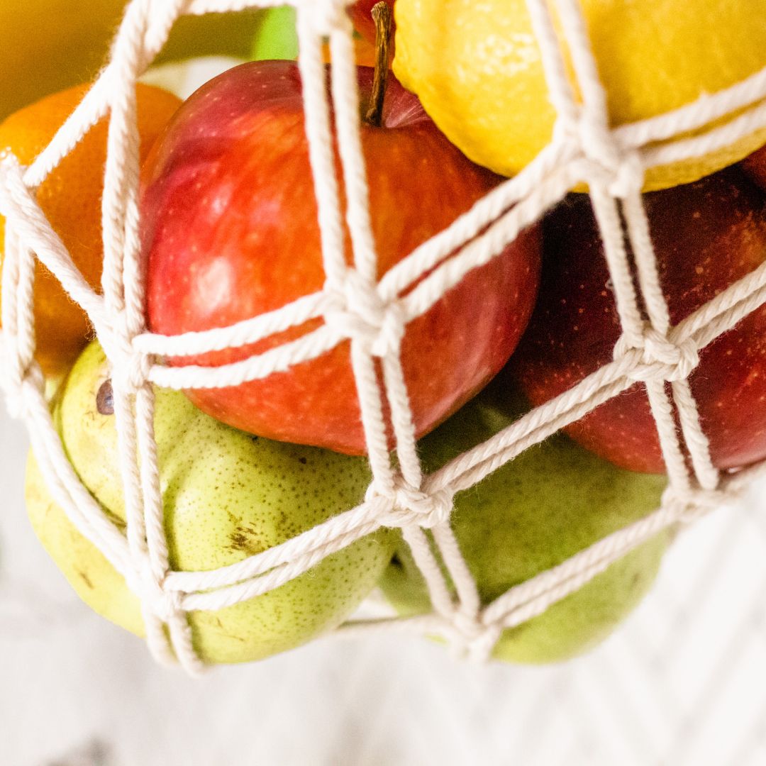 a very close image of a handmade macrame fruit hammock showing the close interlaced cotton holding various fresh fruits in vibrant colours a red apple is clearly seen in this image.